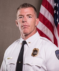 Uniformed police officer poses in front of an American flag indoors.