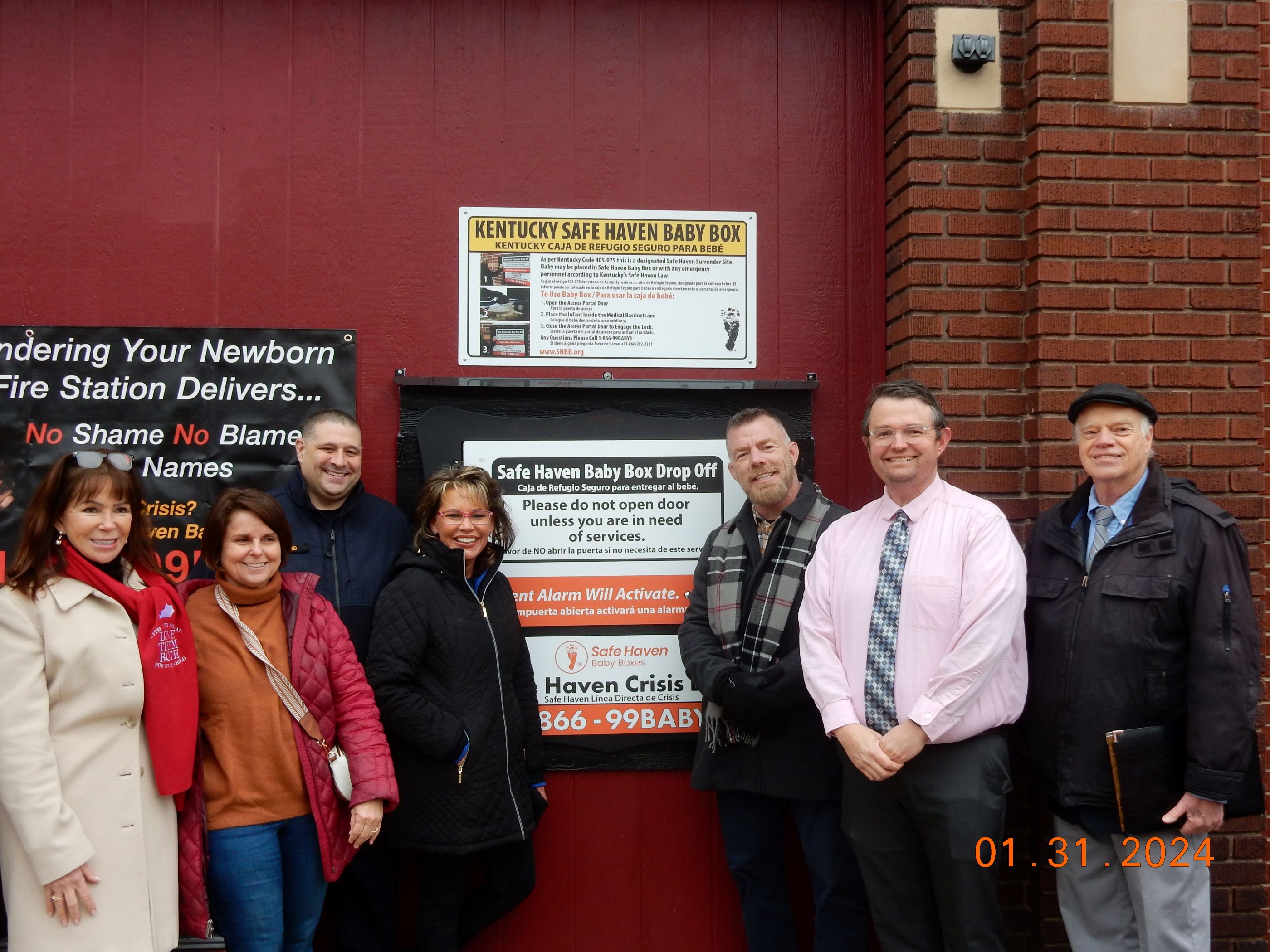 Group of people standing in front of a Safe Haven Baby Box sign on a brick wall.