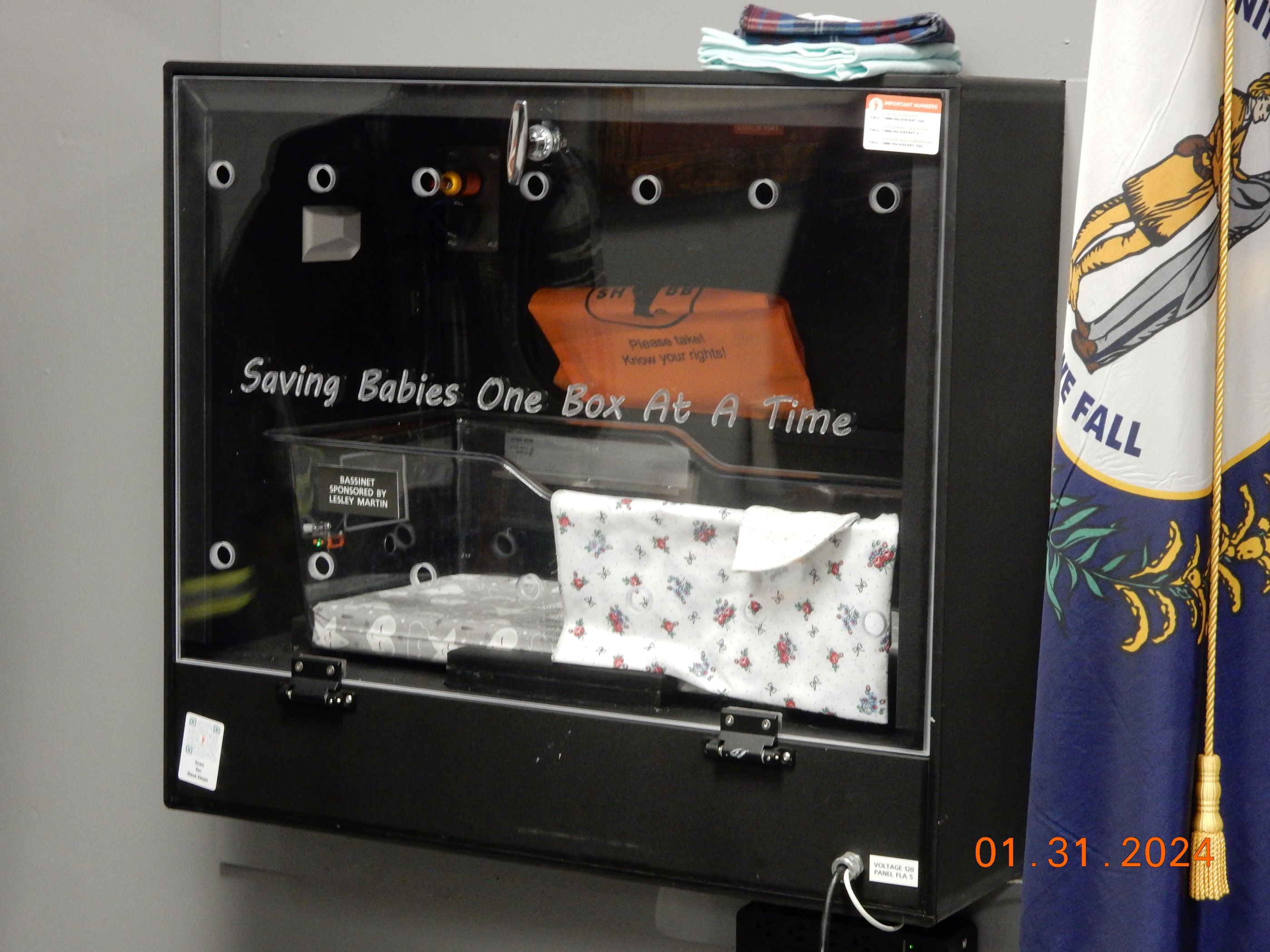 Safe Haven Baby Box installed in a wall with a flag adjacent, featuring a transparent front and blankets inside.