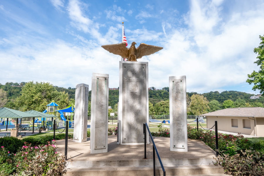 Monument with an eagle and American flag on top, surrounded by inscribed pillars, situated in a park with a playground nearby.