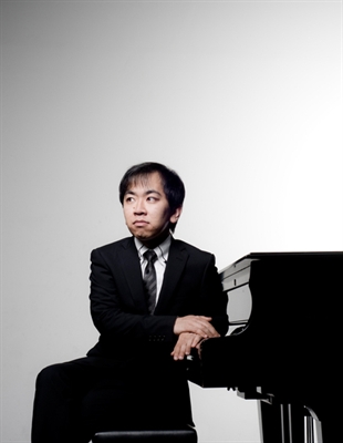 A person in a suit sits on a stool next to a grand piano against a plain background.