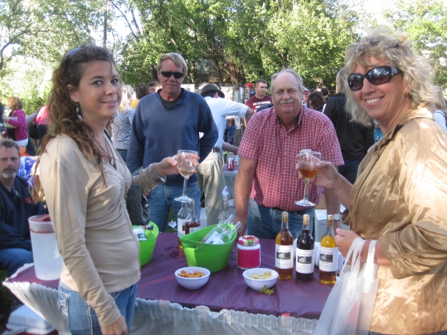 People gather at an outdoor event with a table displaying wine bottles and snacks under a canopy of trees.