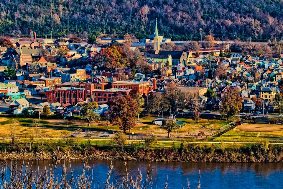 Aerial view of a town with residential buildings, a church, and a school near a riverbank, surrounded by wooded hills.