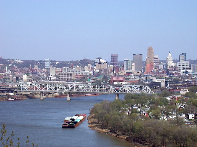 Aerial view of a river with a barge, a bridge, and a city skyline in the background.