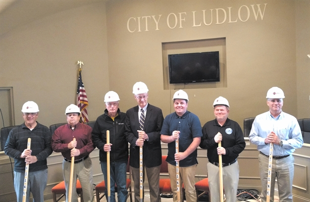 Seven individuals in hard hats holding shovels stand in front of a sign reading 