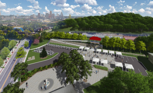 Aerial view of a landscaped area with a parking lot, pavilions, and a fountain, surrounded by trees and overlooking a city skyline.
