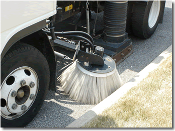 A street sweeper truck in operation, with a rotating brush cleaning the edge of a road beside a grassy curb.