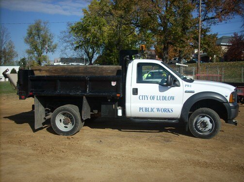 White City of Ludlow Public Works dump truck parked on a dirt lot, viewed from the side, with a black dump bed and trees in the background.