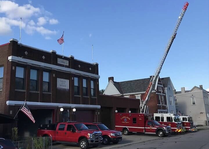 Ludlow firehouse building with American flags displayed, red fire department vehicles parked in front, and a ladder truck with its ladder extended upward beside the building on a clear day.
