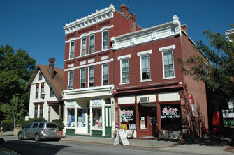Historic brick commercial buildings along a small-town street, with storefront windows, parked cars, and neighboring houses visible in daylight.