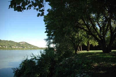 Scenic view of a river bordered by trees and greenery, with a grassy riverbank in the foreground and hills visible across the water under a clear sky.