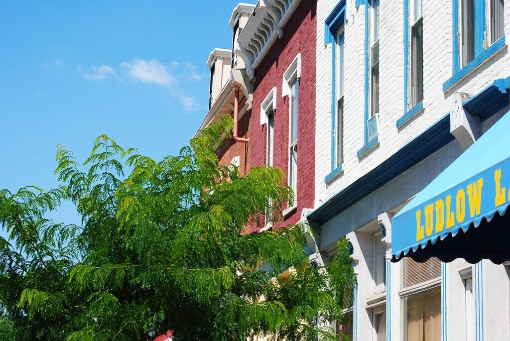 Corner view of a historic commercial block with brick buildings, storefront windows, and light traffic on a sunny day.