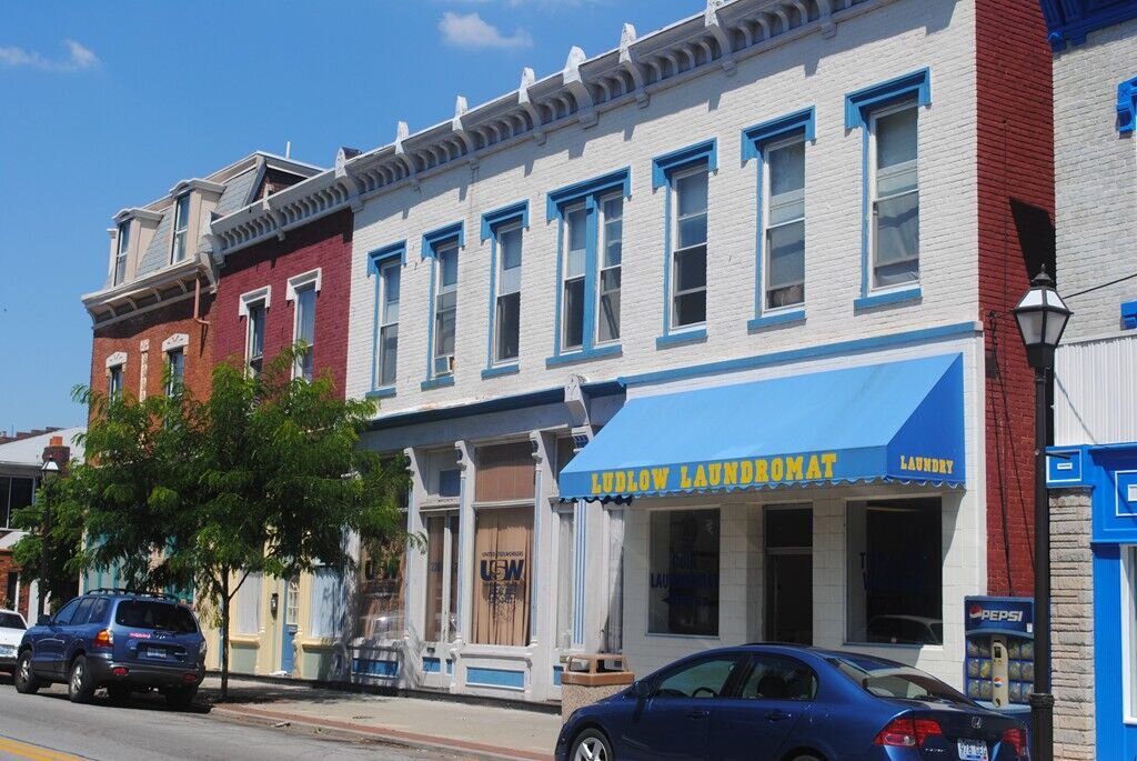 Downtown street lined with brick buildings, parked vehicles, trees, and traffic signals in the distance.