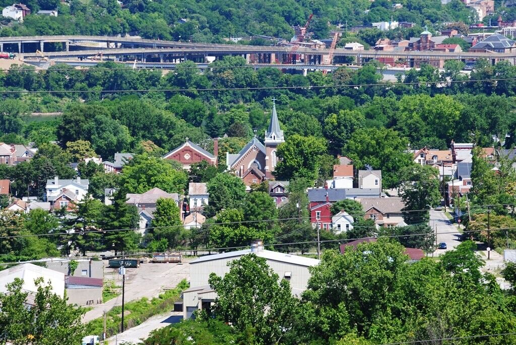 Elevated view of a town with houses, churches, and trees, with a highway and construction visible on a hillside in the background.