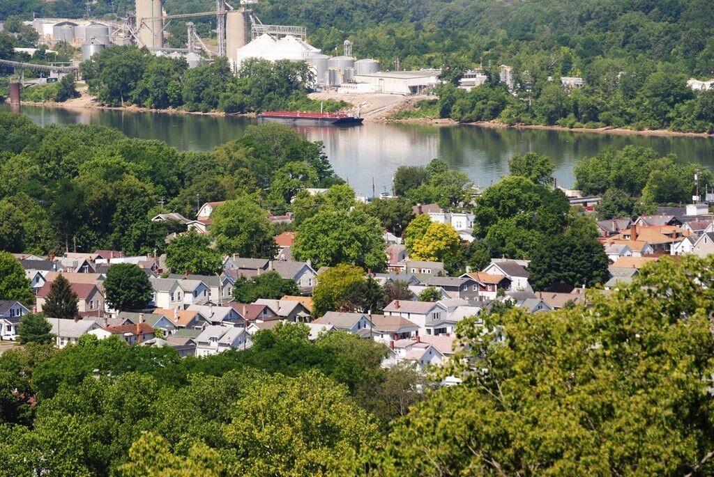 Aerial view of a residential neighborhood with tree-lined streets beside a river, with industrial buildings visible across the water.