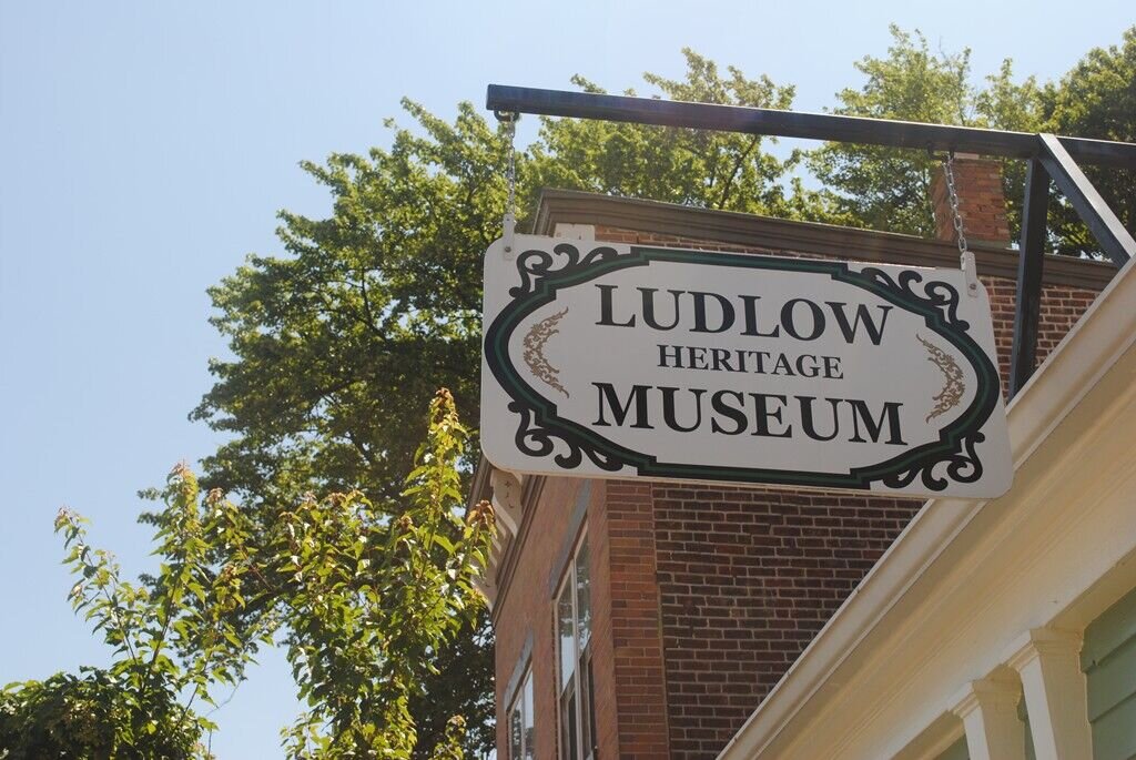 Hanging sign reading ‘Ludlow Heritage Museum’ mounted outside a brick building with trees in the background.