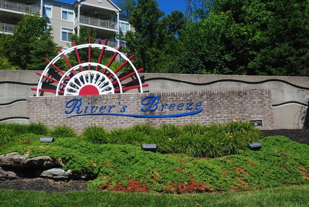 Landscaped entrance sign reading ‘River’s Breeze,’ featuring a decorative wheel design and greenery in front of apartment buildings.