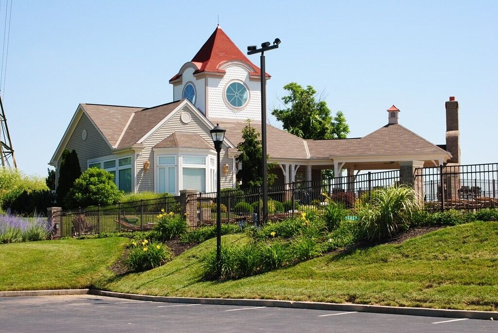 Community clubhouse building with a tower roof, covered patio, black metal fencing, and landscaped grounds on a sunny day.