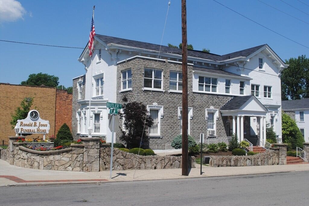 Two-story historic stone and brick building at a street corner with a sign reading ‘Ronald B. Jones Funeral Home’ and an American flag displayed.